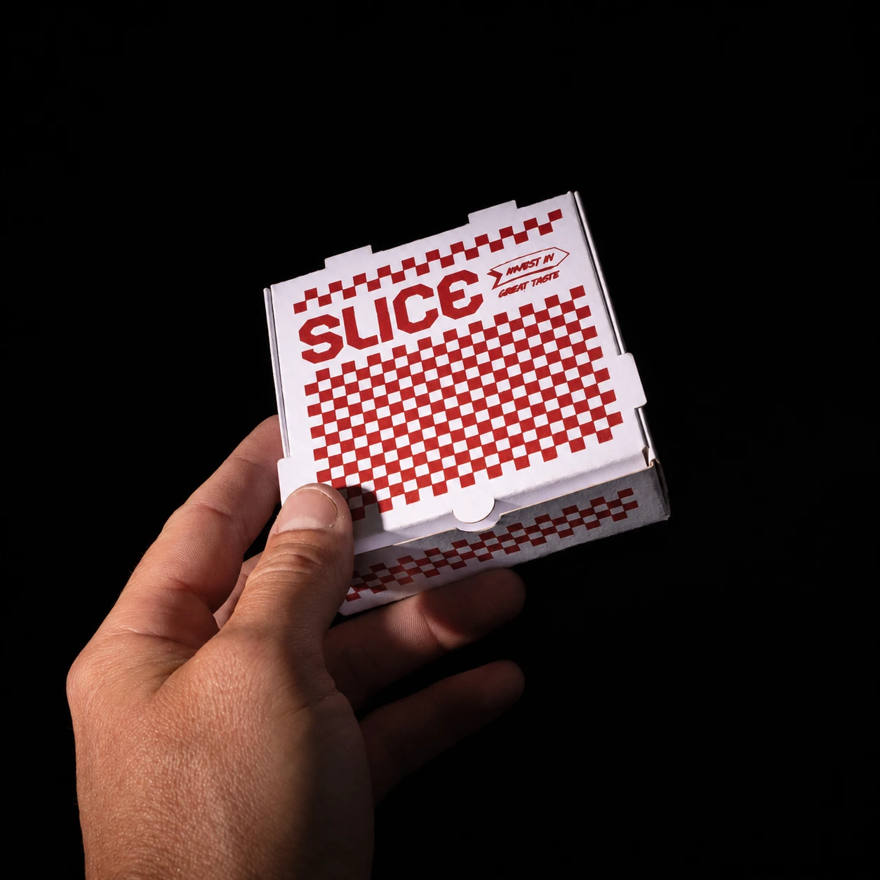 A hand holding the white Slice mini pizza box, showing its scale and red silk-screened checkerboard graphics against black.
